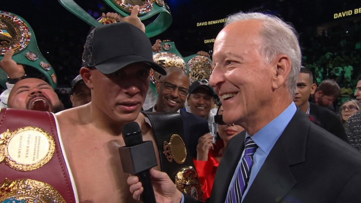 David Benavidez celebrates a dominant victory over Gilberto Ramirez at T-Mobile Arena, holding multiple WBC belts while giving a post-fight interview, with his team cheering in the background.