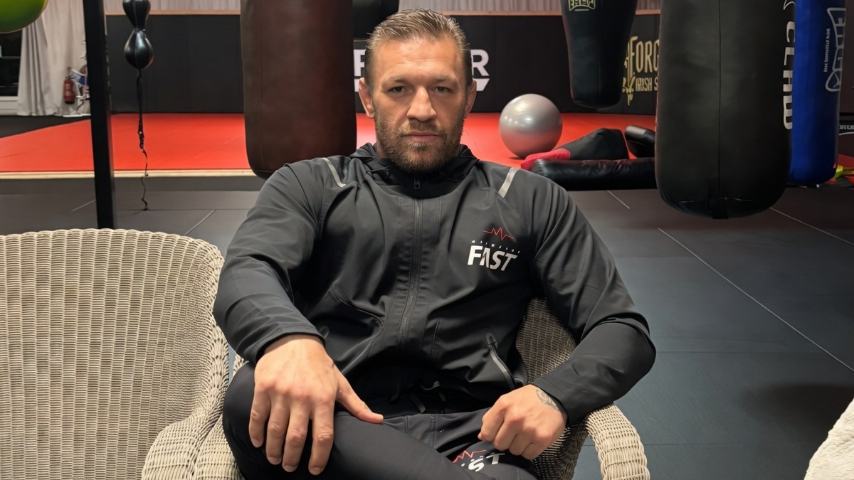 Conor McGregor sits inside a training gym wearing a black tracksuit as he prepares for his return to competition following a lengthy injury layoff