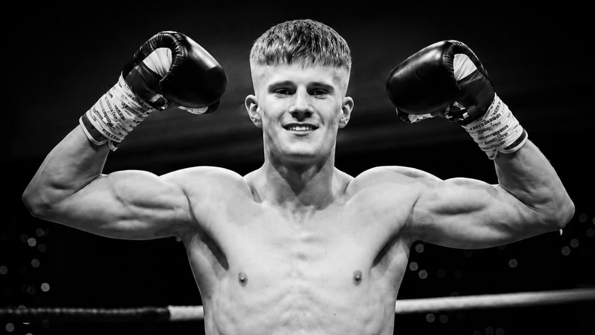 Black-and-white portrait of boxer Colin Cairney posing with raised gloves in a boxing ring