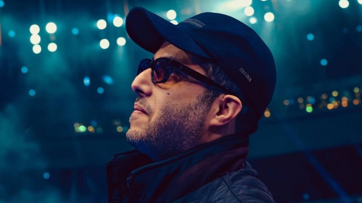 Turki Alalshikh wearing a black cap and sunglasses while looking toward arena lights at a boxing event
