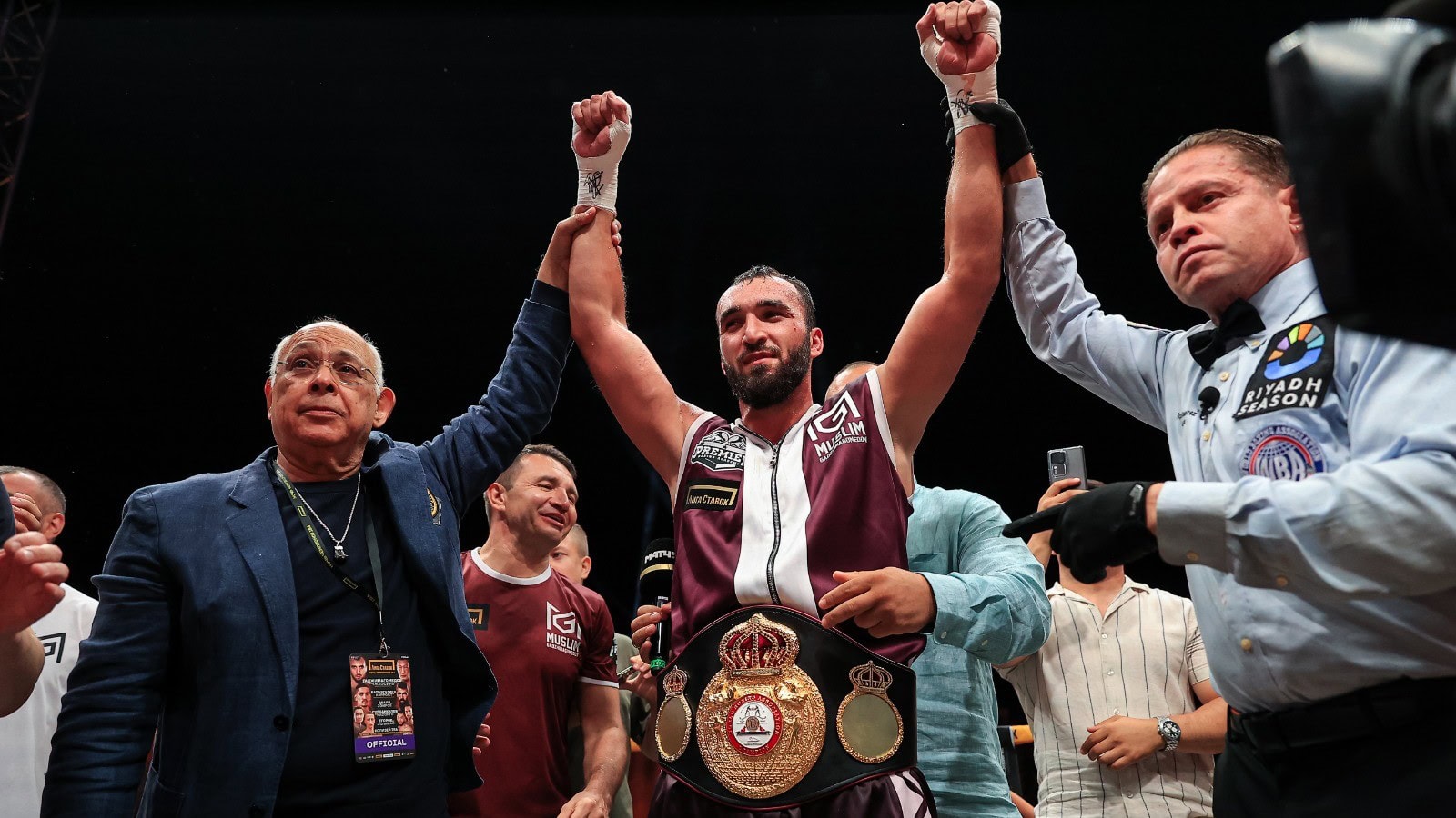Muslim Gadzhimagomedov stands in a boxing ring holding the WBA bridgerweight championship belt as officials raise his arms
