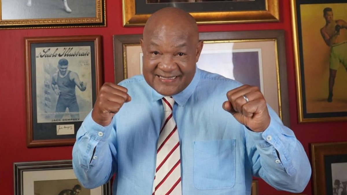 George Foreman raises both fists while smiling in front of framed boxing memorabilia at his home